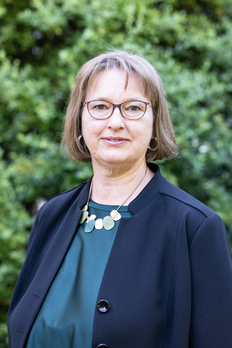 Portrait der Staatssekretärin Frauke Patzke. Sie hat kinnlanges blonde Haare, trägt eine runde Brille, einen baluen Blaser, ein blau-grünes Oberteil und eine Kette mit goldenen und grünen Anhängern. Im Hintergrund sind die Blätter eines Baums.