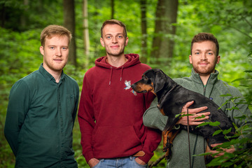 Jan, Simon und Felix (mit einem Dackel auf dem Arm) vom Portal www.forsterklaert.de stehen im Wald.