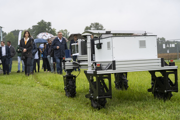 Ein Robotor fährt auf einer Wiese, dahinter geht eine Personengruppe.