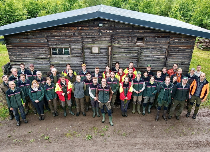 Ein Gruppenfoto zeigt den Prüfungsjahrgang und Prüfungsausschuss 2024 der Länder Nordrhein-Westfalen und Sachsen-Anhalt am Tag der Waldprüfung im Forstbetrieb Ostharz (Sachsen-Anhalt) vor einer Blockhütte