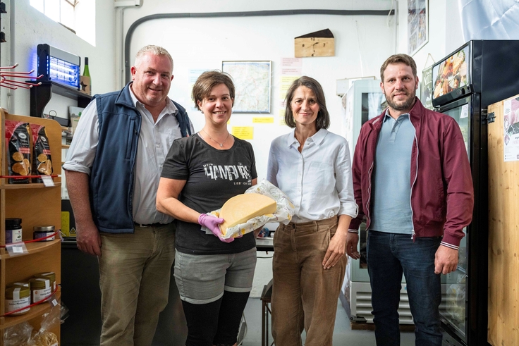 Ministerin Miriam Staudte neben Frau Heidrich, die einen halben Laib Käse in der Hand hält, und zwei Landwirten