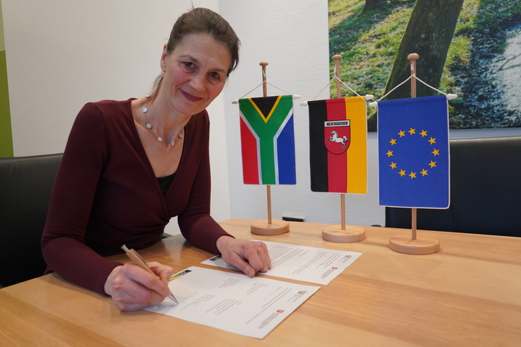 Ministerin Staudte unterzeichnet an einem hellbraunen Tisch in ihrem Bürü die Absichtserklärung