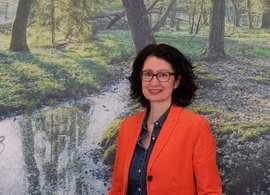 Frau mit lockigen Haaren und roter Jacke vor einem Bild mit Wald