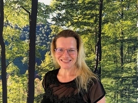 Frau mit blonden Haaren und Schwarzem Oberteil vor einem Bild mit Wald