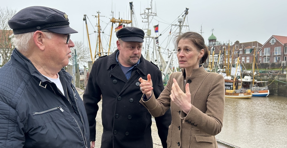 Ministerin Miriam Staudte im Gespräch mit zwei Fischern, im Hintergrund ein Hafen mit Schiffen