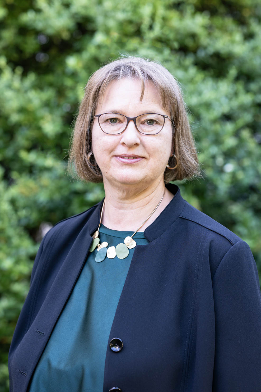 Portrait der Staatssekretärin Frauke Patzke. Sie hat kinnlanges blonde Haare, trägt eine runde Brille, einen baluen Blaser, ein blau-grünes Oberteil und eine Kette mit goldenen und grünen Anhängern. Im Hintergrund sind die Blätter eines Baums.