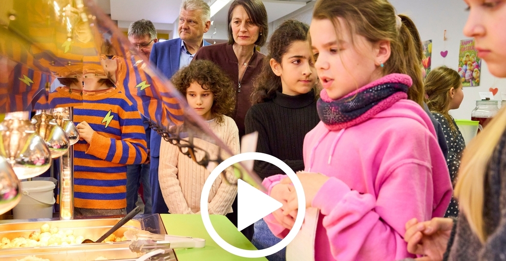 Kinder schauen in einer Kantine auf GN Behälter mit essen. Im Hintergrund Ministerin Miriam Staudte